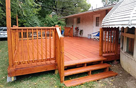 Newly built wooden deck with steps, surrounded by green grass and trees, adjacent to a house with a silver awning.