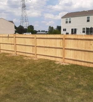 A construction worker builds a wooden fence in a backyard, surrounded by grass and trees.