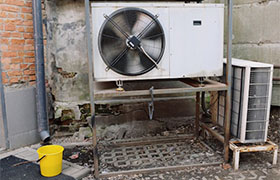 An outdoor air conditioning unit on a metal stand, featuring a large fan, a filter beside it, and a yellow bucket below.