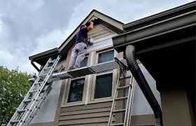 A worker is painting the eaves of a house while standing on a ladder, with cloudy skies in the background.